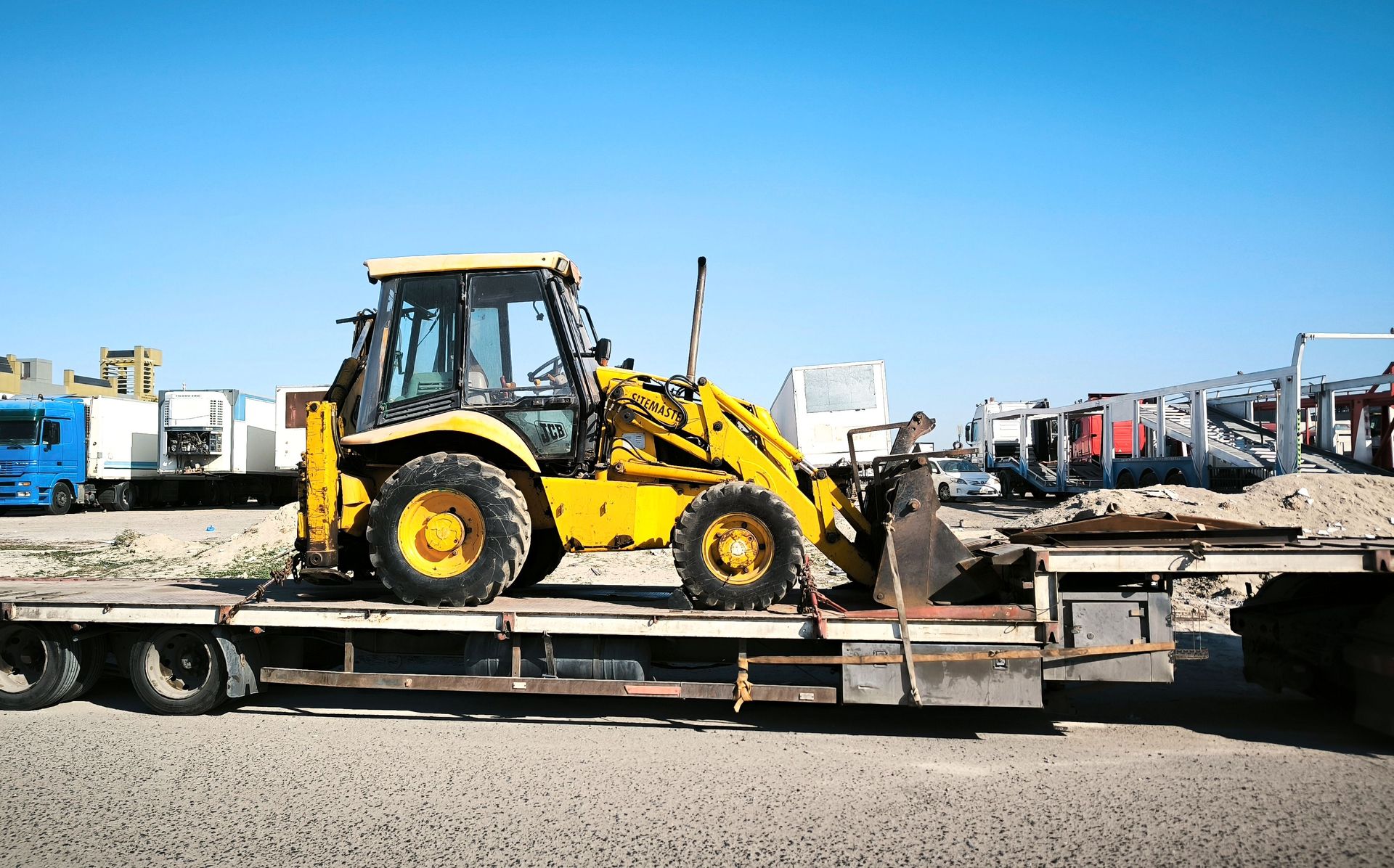 A yellow construction vehicle is seen on a flatbed trailer in an industrial area under a clear blue sky.