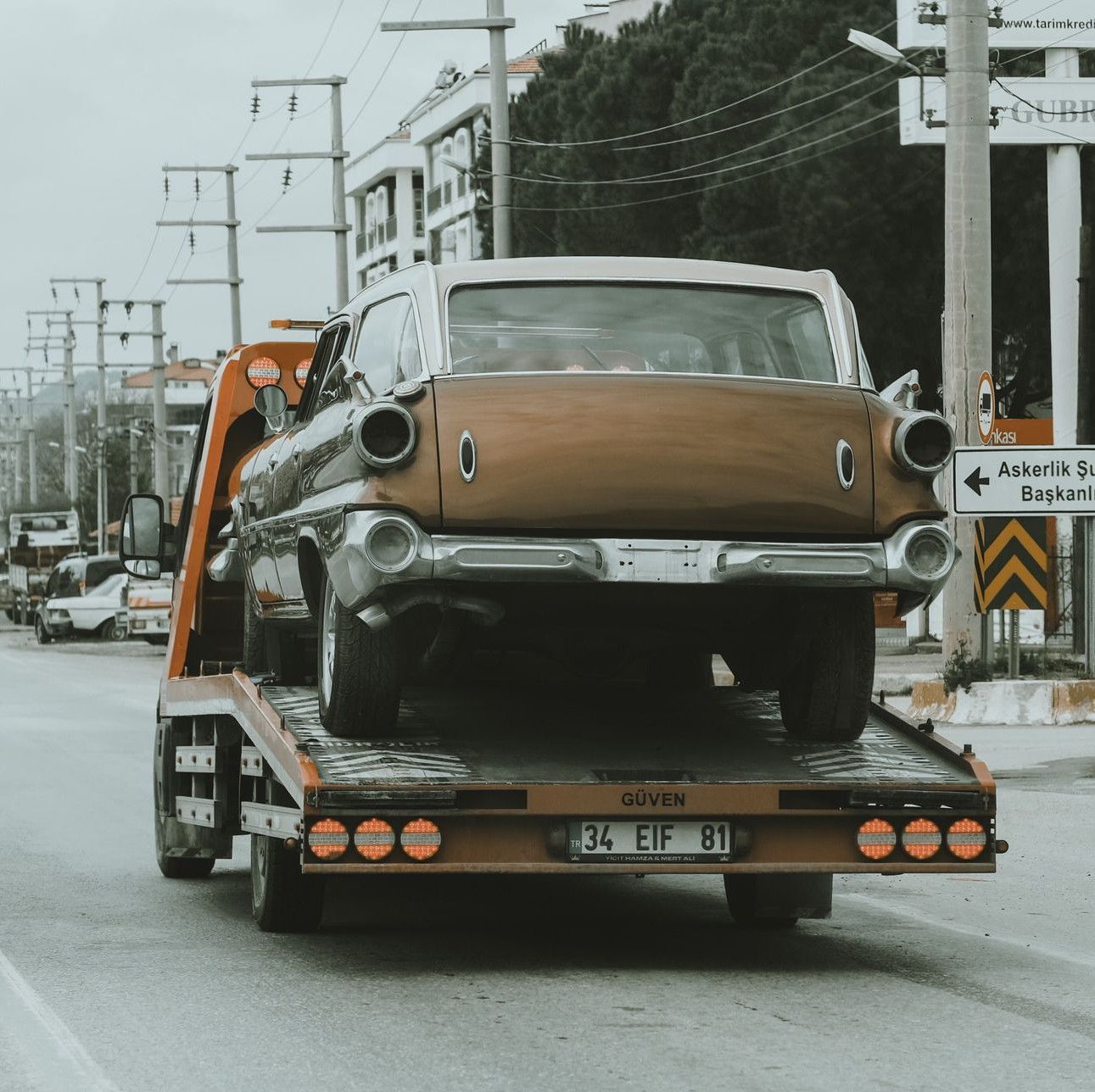 A vintage car is being towed through city streets on a tow truck, showcasing an urban environment.